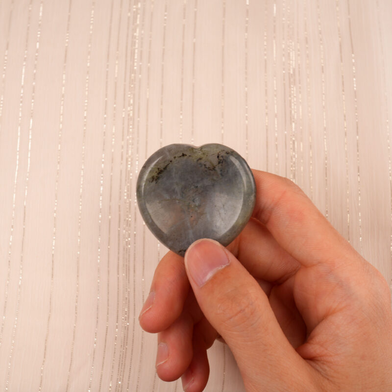 Fingers holding a polished labradorite heart worry stone, highlighting its size and glow