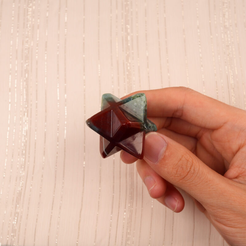 Hand holding a natural Indian Agate Merkaba crystal, showing its compact size and earthy red-green tones under natural light.