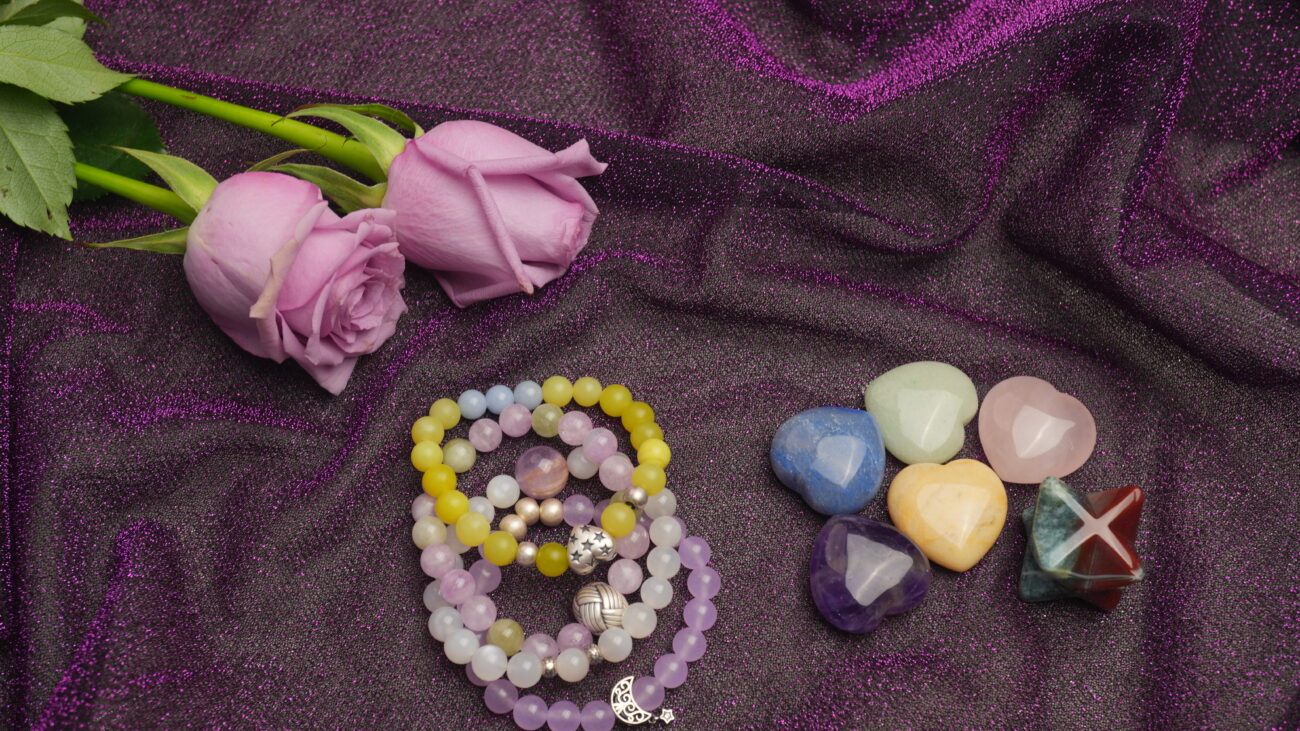 Lavender roses with stacking crystal beaded bracelets and heart-shaped stones on a purple fabric background
