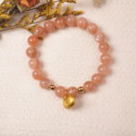 Lifestyle photo of peach moonstone bracelet with gold charm placed on soft fabric with warm light.