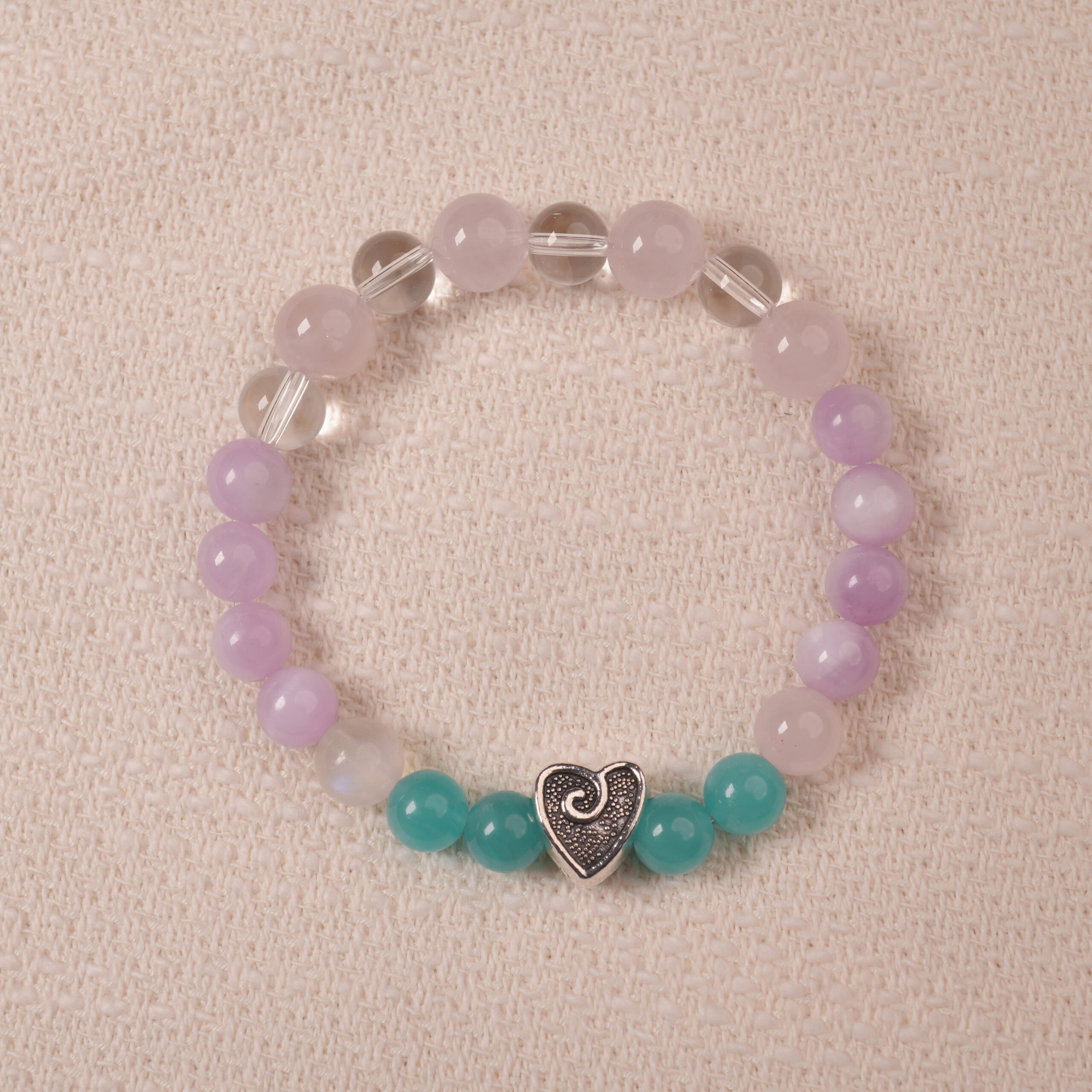 amethyst-prehnite-bracelet-main Amethyst and Prehnite bracelet featuring smooth, polished beads in shades of purple and green, with a central silver heart-shaped bead. The bracelet is displayed on a soft fabric surface, emphasizing its elegance and soothing colors.