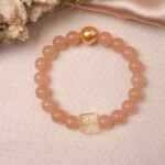 Lifestyle photo of peach moonstone bracelet with gold heart and citrine bead on beige fabric.