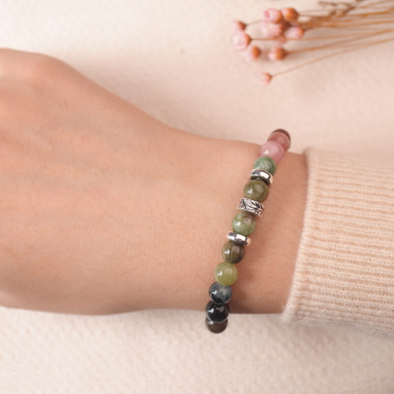 A Tourmaline bracelet laid on a soft fabric surface, with a rich array of colors including pink, green, black, and purple beads, accented by silver spacers. The bracelet is positioned near delicate flowers, adding a gentle, natural touch to the composition.