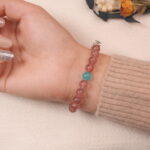 A model wearing the Strawberry Quartz and Angelite bracelet on their wrist. The heart-shaped silver charm adds a touch of elegance and the combination of soothing stones promotes calm and balance.