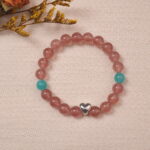 The Strawberry Quartz and Angelite bracelet laid on a soft fabric background, with its golden accents and heart-shaped silver charm shining under the light, creating a warm and inviting atmosphere.