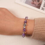 A model wearing the Amethyst and Clear Quartz bracelet on their wrist, showing the front view. The vibrant purple of the amethyst and the transparent clear quartz are beautifully displayed with the silver cross charm at the center.