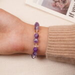 A model wearing the Amethyst and Clear Quartz bracelet on their wrist, showing the back view. The bracelet's smooth beads and the subtle silver cross accent are clearly visible.
