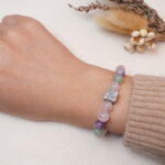 A wrist wearing a bracelet featuring rose quartz, amethyst, green aventurine, and a silver rose charm.