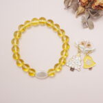 A yellow quartz bead bracelet with a pearl center and a pair of festive yellow bell and Christmas tree earrings set beside dried leaves.