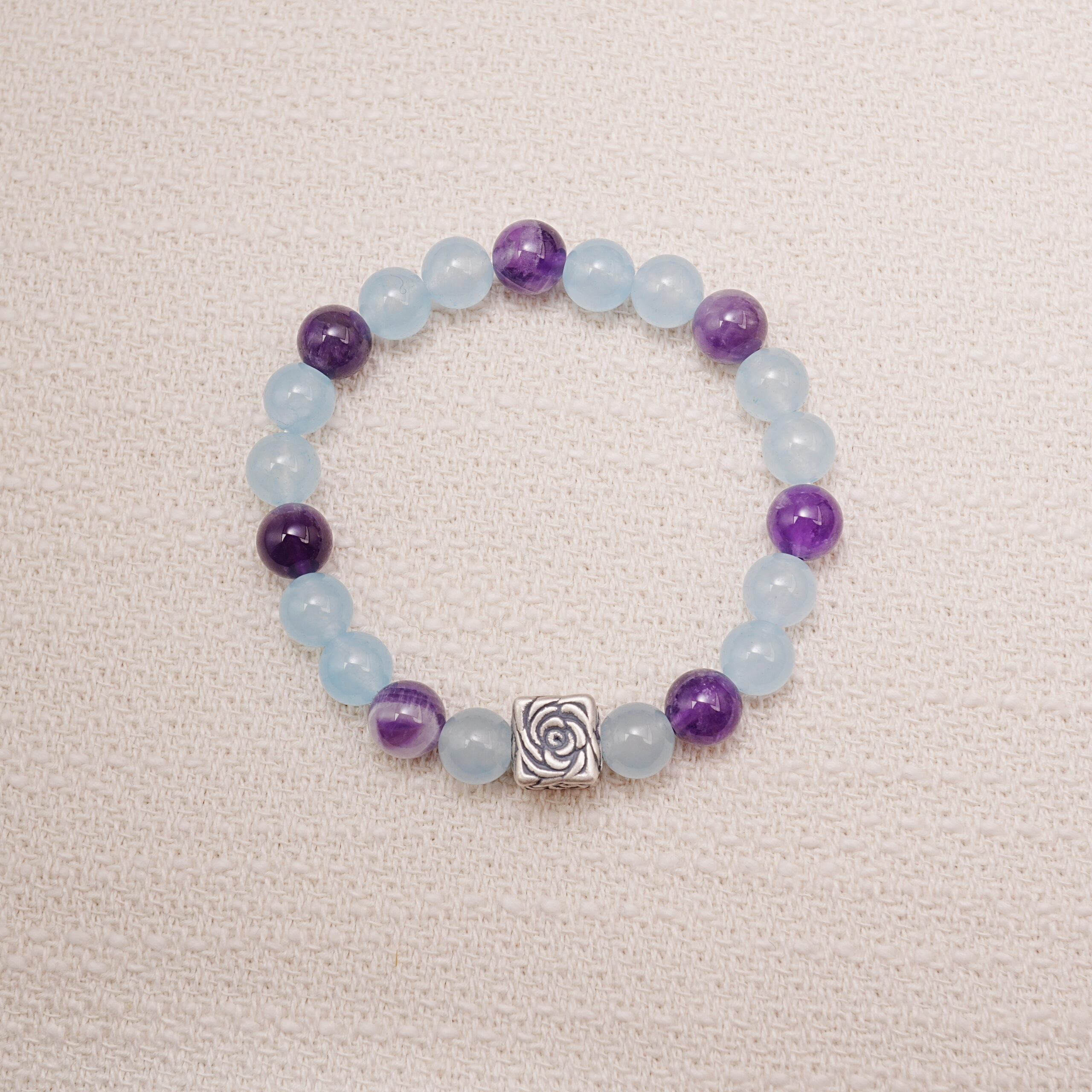 aquamarine-amethyst-rose-bead-bracelet-main Aquamarine and amethyst beaded bracelet with a silver rose charm displayed on a soft fabric background.