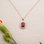 Rose gold ruby halo pendant necklace styled with flowers on a textured beige surface.
