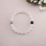 Flat-lay image of a moonstone bracelet featuring one black obsidian bead and a silver accent on a textured beige background.