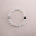 White moonstone beaded bracelet with a single black obsidian accent bead, displayed on a soft beige background.
