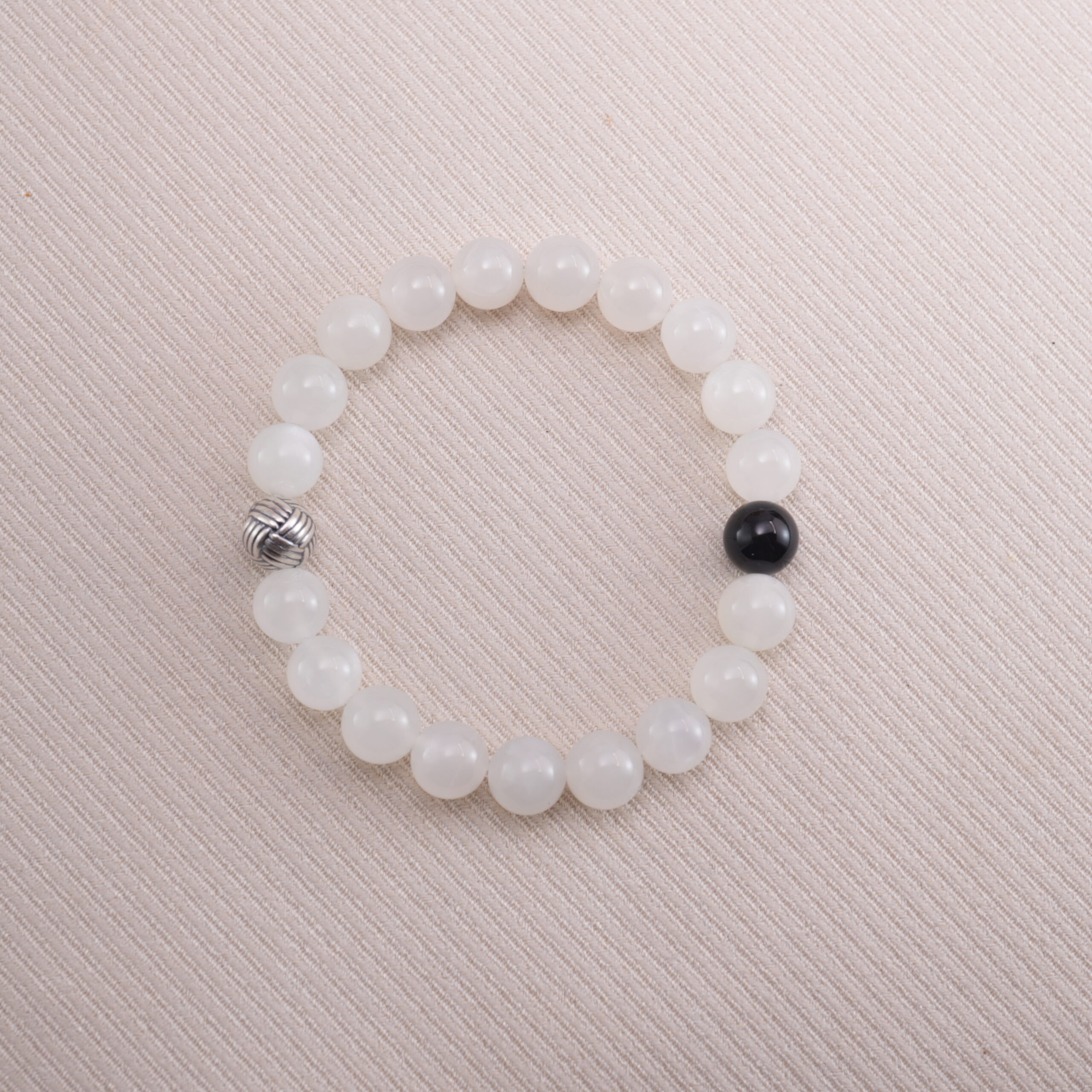 moonstone-obsidian-bracelet-main White moonstone beaded bracelet with a single black obsidian accent bead, displayed on a soft beige background.