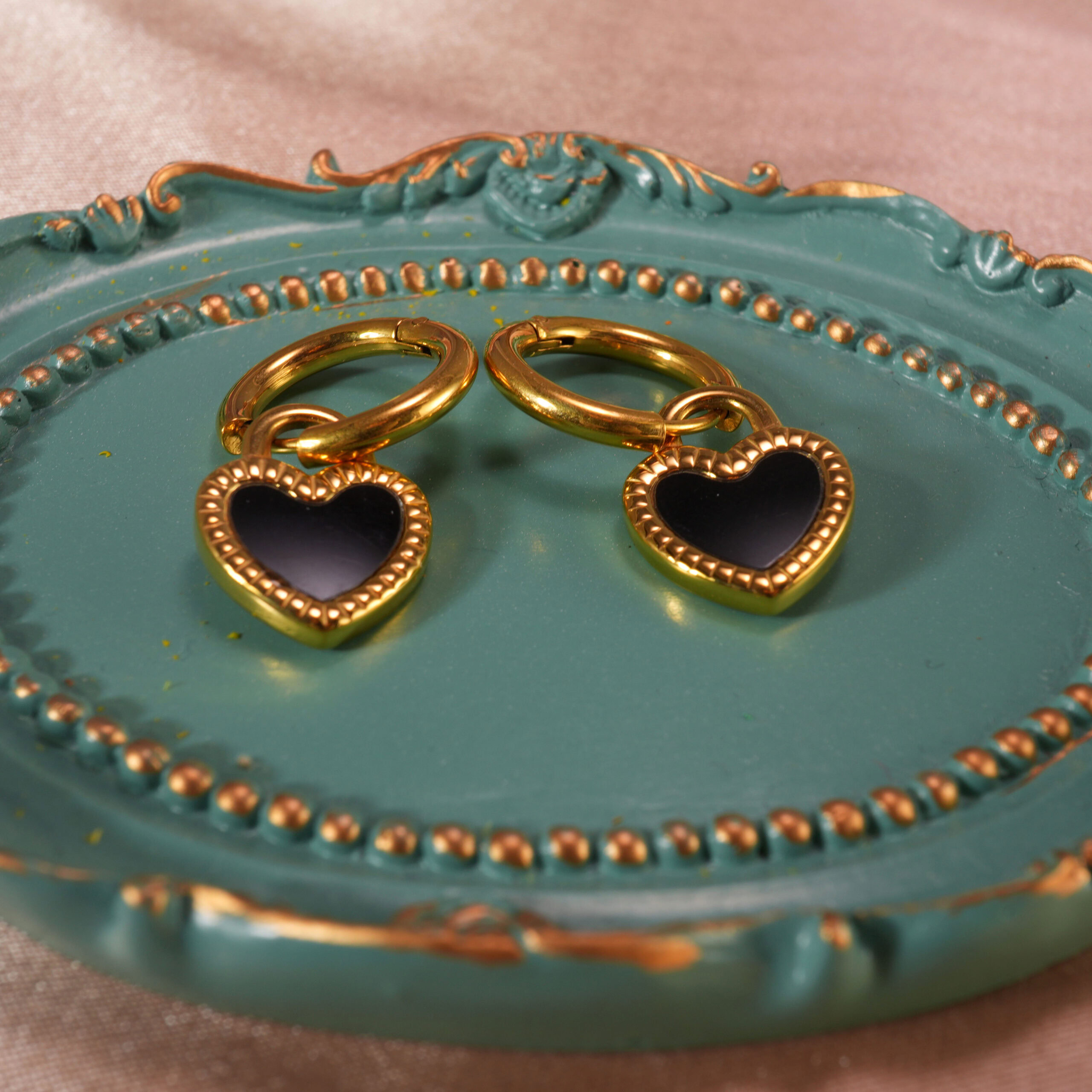 heart-enamel-hoop-earrings-black-main.jpg Heart-shaped black enamel hoop earrings displayed on a decorative teal tray