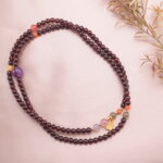 A long garnet beaded necklace with amethyst, yellow crystal, and red agate accents styled beside decorative flowers.