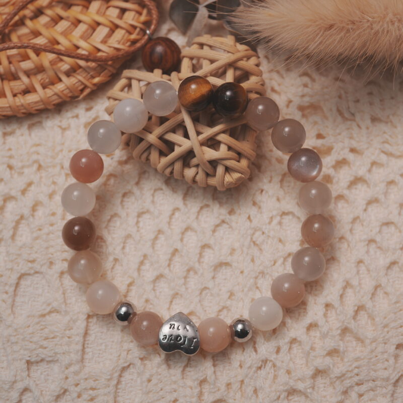 Rainbow moonstone and tiger’s eye bracelet with a silver “I love you” heart accent displayed in a natural lifestyle setting under daylight, capturing the balance between glowing moonstone and warm tiger’s eye tones.